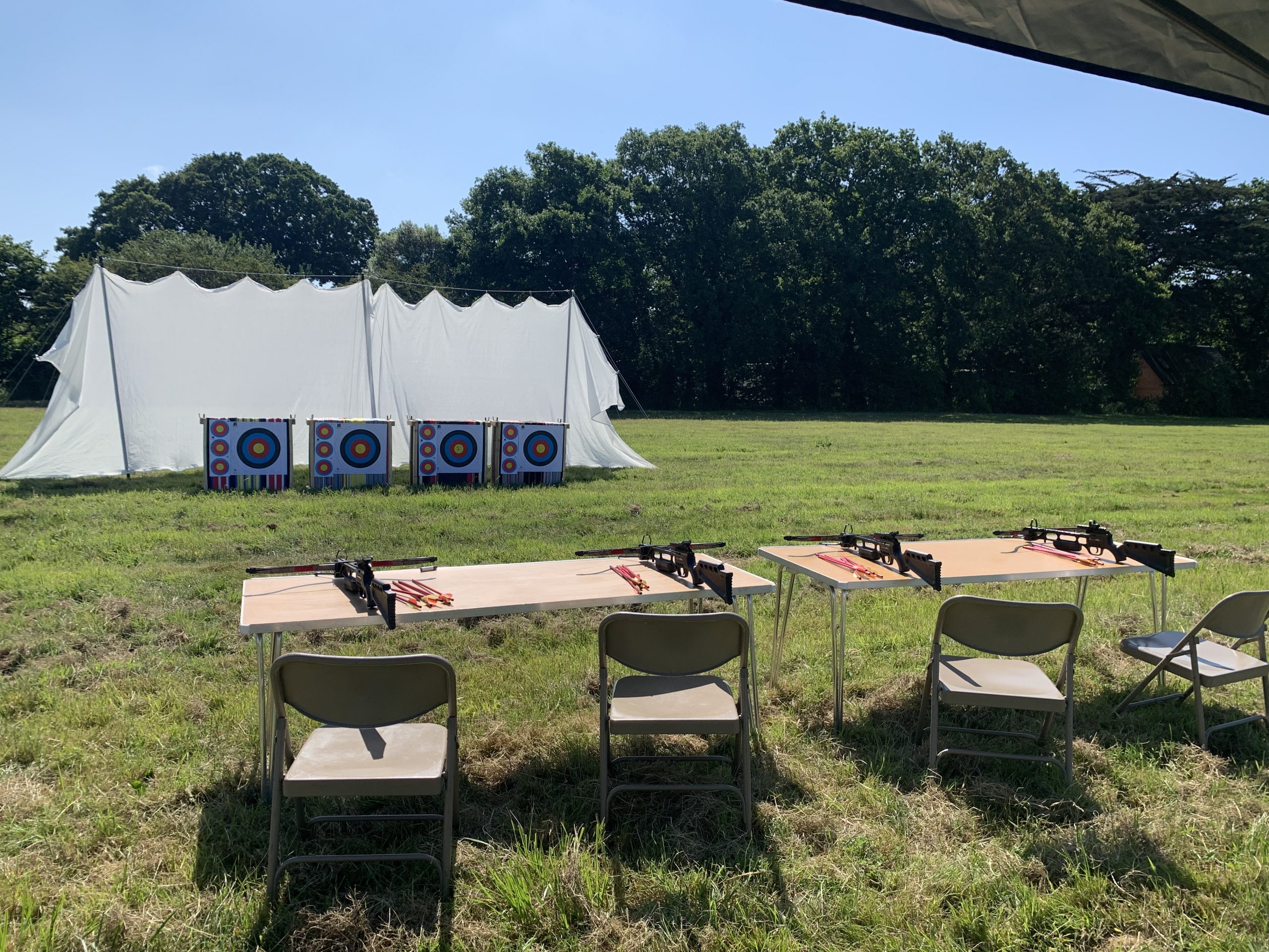 Crossbow range with 4 crossbows on tables in a field with a large net and 4 targets.
